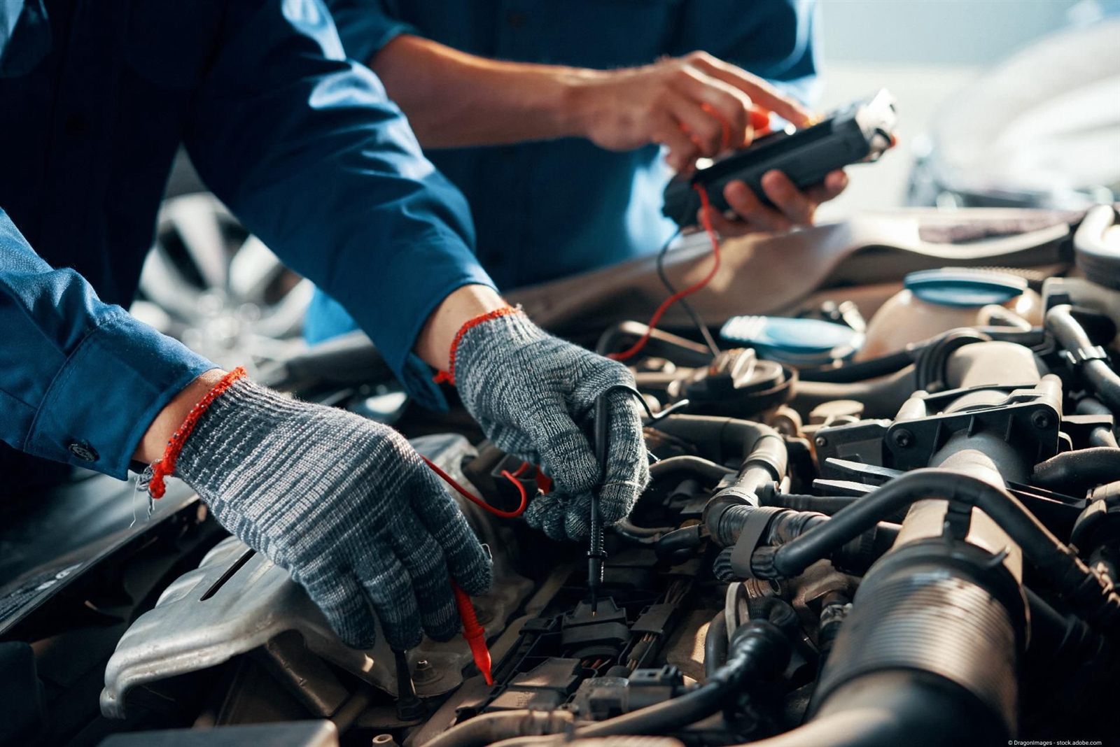 Zwei Mechaniker in Arbeitshandschuhen messen mit einem Messgerät an Steckverbindungen im Motorraum eines Autos
